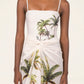 A woman wears the Lavanda Arena Pareo by Agua By Agua Bendita—a white dress with thin straps, tropical palm tree print, and a sarong-style knot at the waist—ideal as chic beachwear or a stylish swimsuit cover-up.