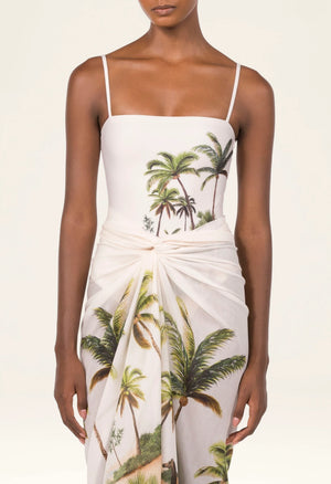 A woman wears the Lavanda Arena Pareo by Agua By Agua Bendita—a white dress with thin straps, tropical palm tree print, and a sarong-style knot at the waist—ideal as chic beachwear or a stylish swimsuit cover-up.