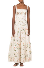 A woman wears the Lima Navegante Maxi Dress by Agua By Agua Bendita, a sleeveless, ankle-length linen dress with a floral pattern, square neckline, and hand-embroidered straps, paired with white heeled sandals.