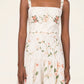 A woman wears the Lima Navegante Maxi Dress by Agua By Agua Bendita— a sleeveless, white linen dress with a floral print and hand-embroidered straps— as she stands against a plain background.
