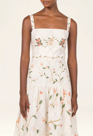 A woman wears the Lima Navegante Maxi Dress by Agua By Agua Bendita— a sleeveless, white linen dress with a floral print and hand-embroidered straps— as she stands against a plain background.