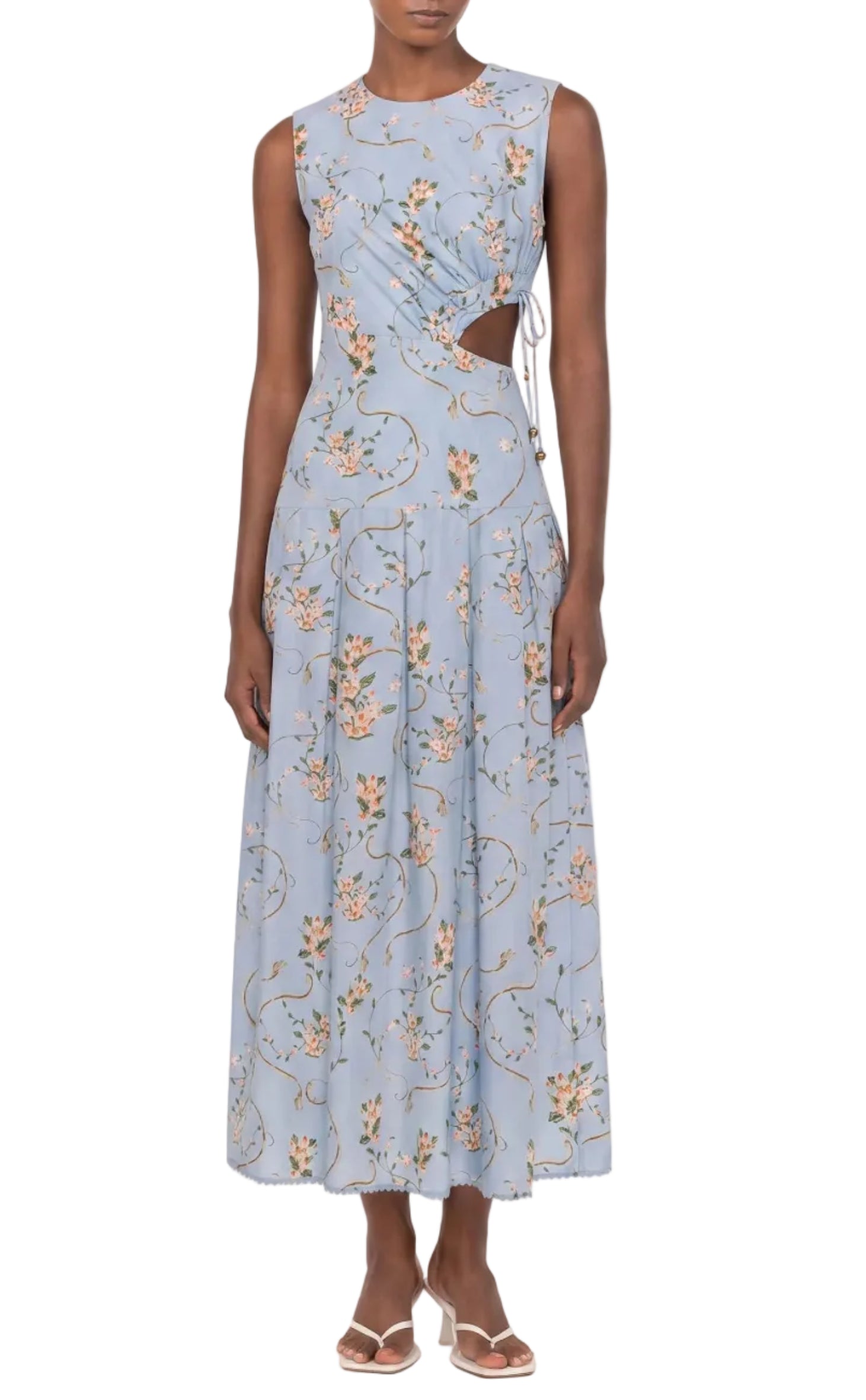 A person wearing the Otono Navegante Midi Dress by Agua By Agua Bendita—a sleeveless, light blue floral dress with side cutouts—paired with white sandals.