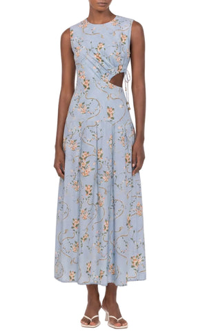 A person wearing the Otono Navegante Midi Dress by Agua By Agua Bendita—a sleeveless, light blue floral dress with side cutouts—paired with white sandals.