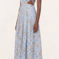 A woman wears the Otono Navegante Midi Dress by Agua By Agua Bendita, a sleeveless, light blue floral dress with side cutouts, paired with flat sandals, standing against a plain background.
