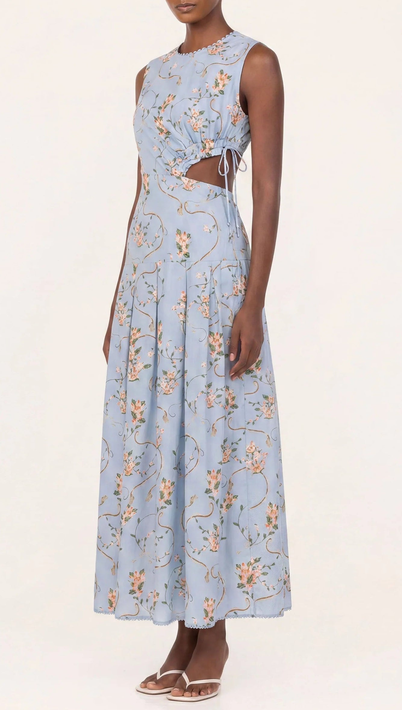 A woman wears the Otono Navegante Midi Dress by Agua By Agua Bendita, a sleeveless, light blue floral dress with side cutouts, paired with flat sandals, standing against a plain background.