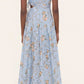 A woman is seen from the back wearing the Otono Navegante Midi Dress by Agua By Agua Bendita, a sleeveless, light blue floral dress with a fitted waist and A-line skirt, paired with white heeled sandals.
