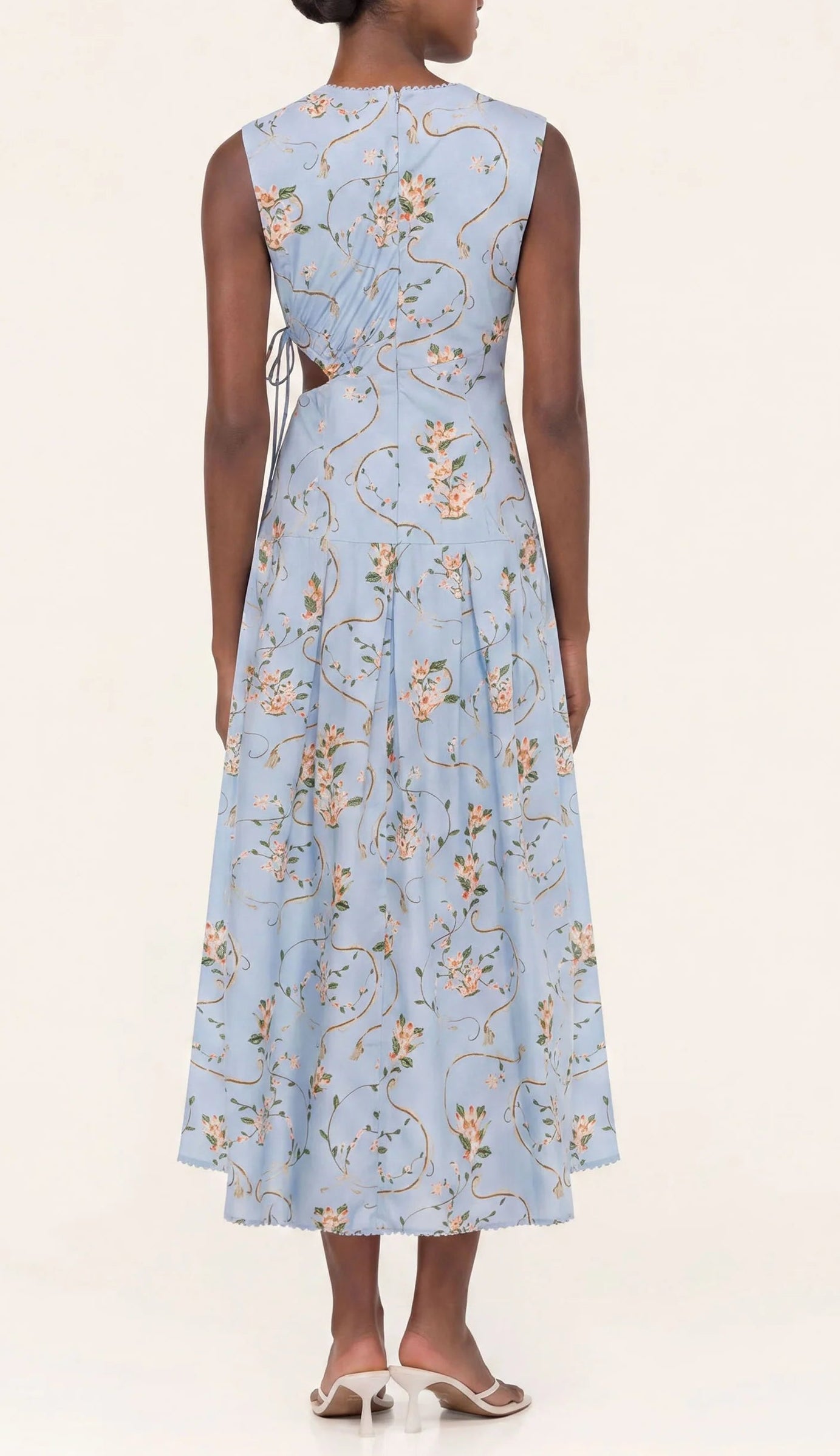A woman is seen from the back wearing the Otono Navegante Midi Dress by Agua By Agua Bendita, a sleeveless, light blue floral dress with a fitted waist and A-line skirt, paired with white heeled sandals.