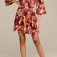 A woman models the Figue Izzie Dress, an A-line band collar design with long sleeves and a tie waist, showcasing maroon, pink, and yellow patterns. She pairs it with black strappy heels.