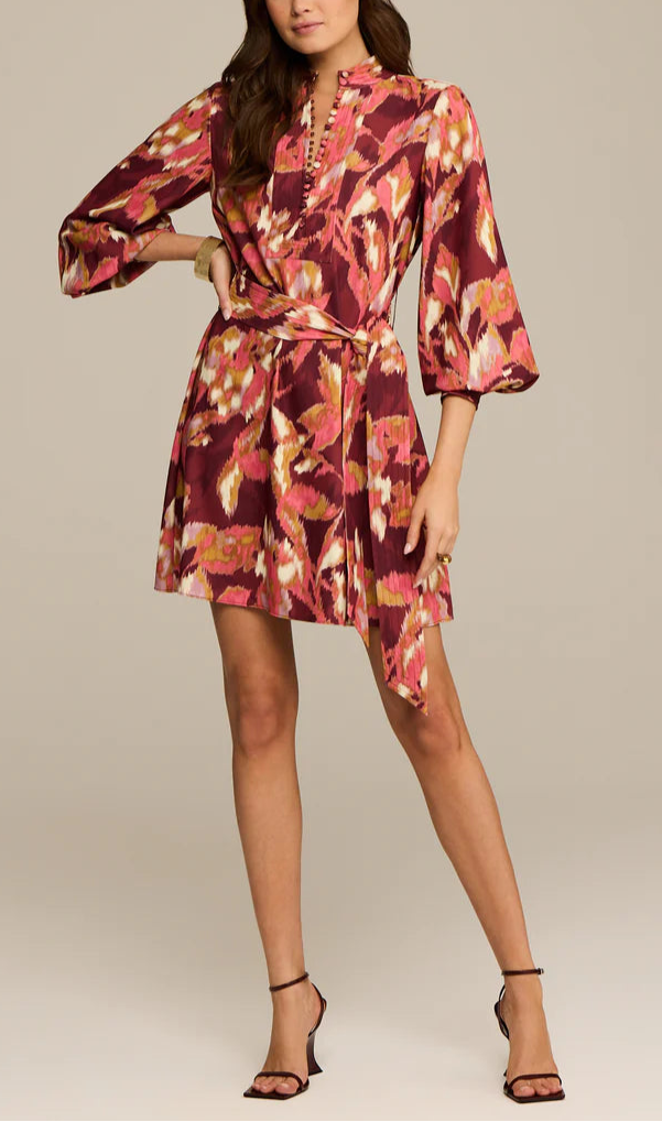 A woman models the Figue Izzie Dress, an A-line band collar design with long sleeves and a tie waist, showcasing maroon, pink, and yellow patterns. She pairs it with black strappy heels.