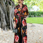 A woman wearing sunglasses, a flowing floral blouse, and Farm Rio's Chenille Flowers Black Pants stands on gravel near a large tree, holding a small handbag with greenery in the background.