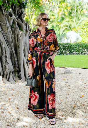A woman wearing sunglasses, a flowing floral blouse, and Farm Rio's Chenille Flowers Black Pants stands on gravel near a large tree, holding a small handbag with greenery in the background.