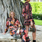 Two women in matching FARM Rio outfits pose by a large tree—one sits on a stone bench, the other stands with a small handbag—both highlighting eco-conscious viscose and Farm Rio’s Chenille Flowers Black Pants.