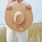 A person in white stands outdoors amid tall grass and sky, holding the Mar Y Sol Jane Hat—a wide-brimmed raffia sun hat—in front of them.