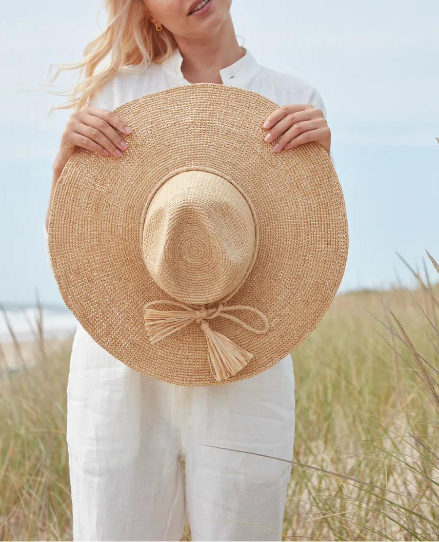 A person in white stands outdoors amid tall grass and sky, holding the Mar Y Sol Jane Hat—a wide-brimmed raffia sun hat—in front of them.