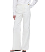 A person wearing Callas Milano's Jane Pant in white organic cotton with an a-line wide-leg, styled with a white long-sleeve shirt and black sandals, stands against a plain background.