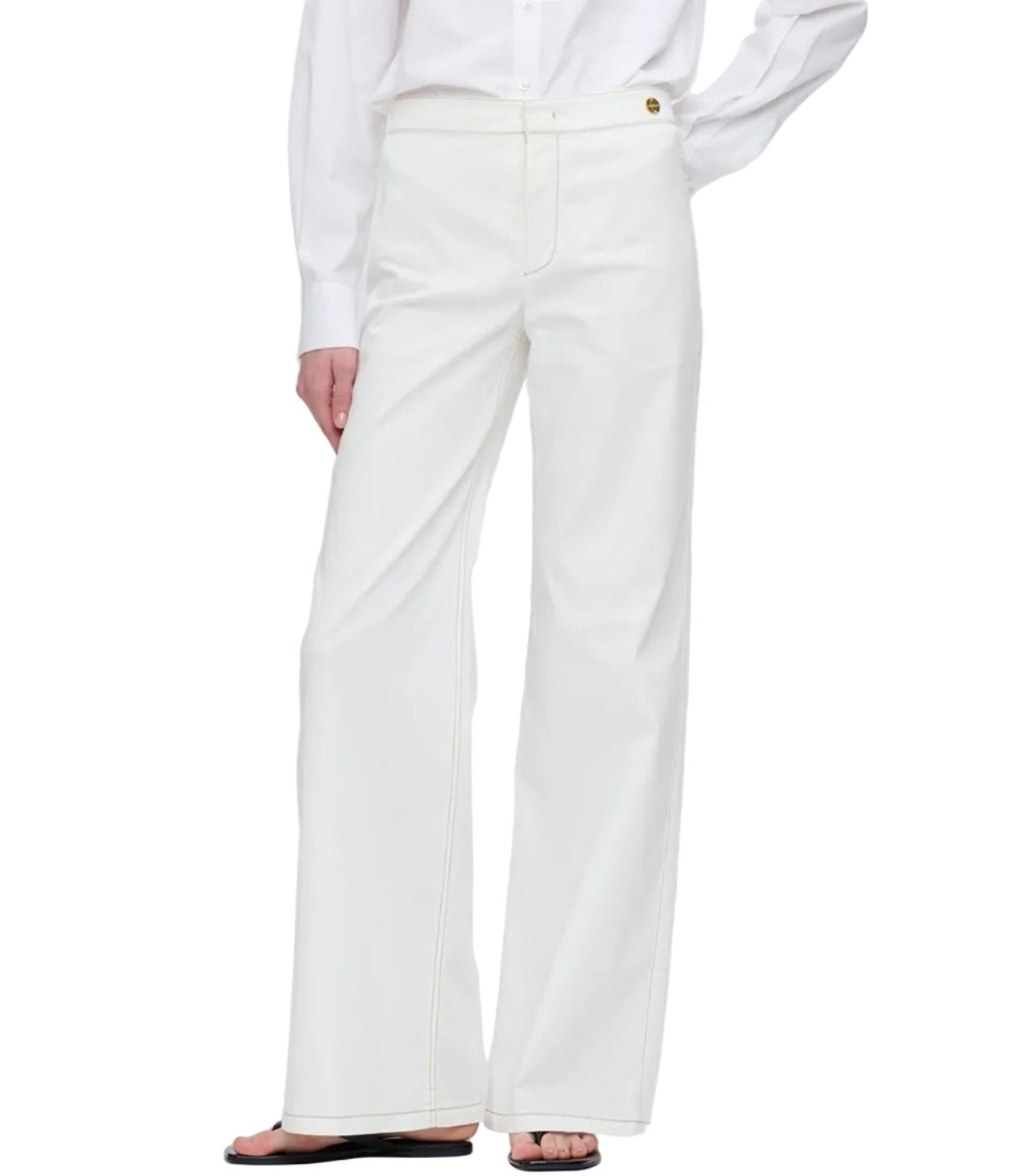 A person wearing Callas Milano's Jane Pant in white organic cotton with an a-line wide-leg, styled with a white long-sleeve shirt and black sandals, stands against a plain background.