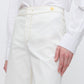 A person wears the Callas Milano Jane Pant—high-rise organic cotton with light yellow stitching, gold button, and A-line flare leg—paired with a white button-up shirt, standing against a plain background.