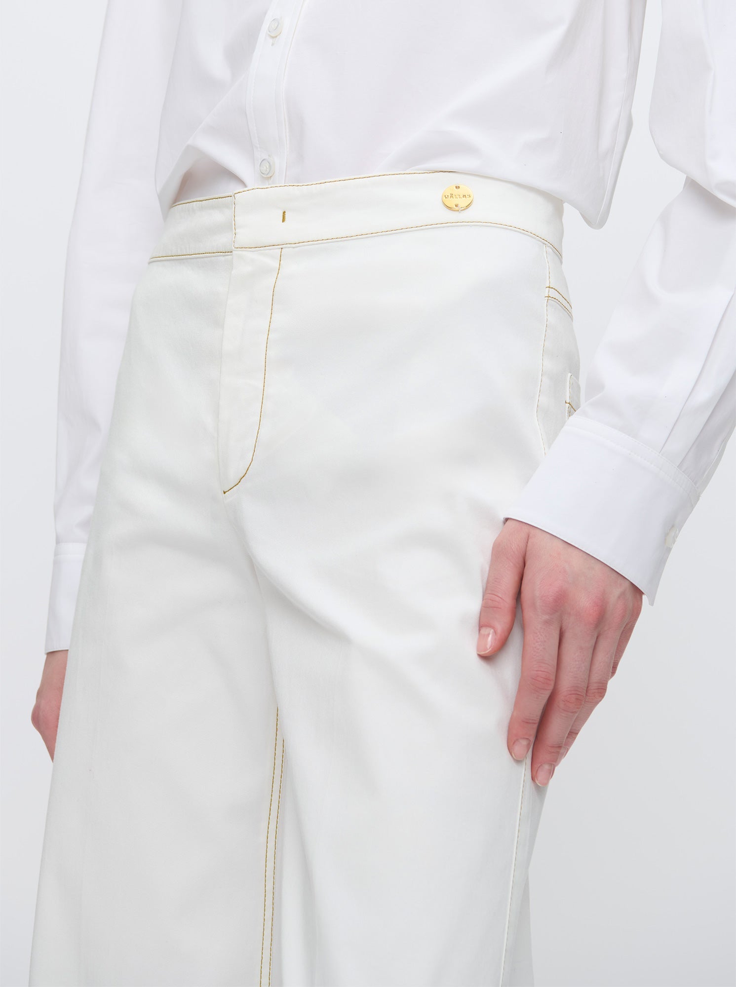 A person wears the Callas Milano Jane Pant—high-rise organic cotton with light yellow stitching, gold button, and A-line flare leg—paired with a white button-up shirt, standing against a plain background.