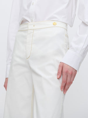 A person wears the Callas Milano Jane Pant—high-rise organic cotton with light yellow stitching, gold button, and A-line flare leg—paired with a white button-up shirt, standing against a plain background.