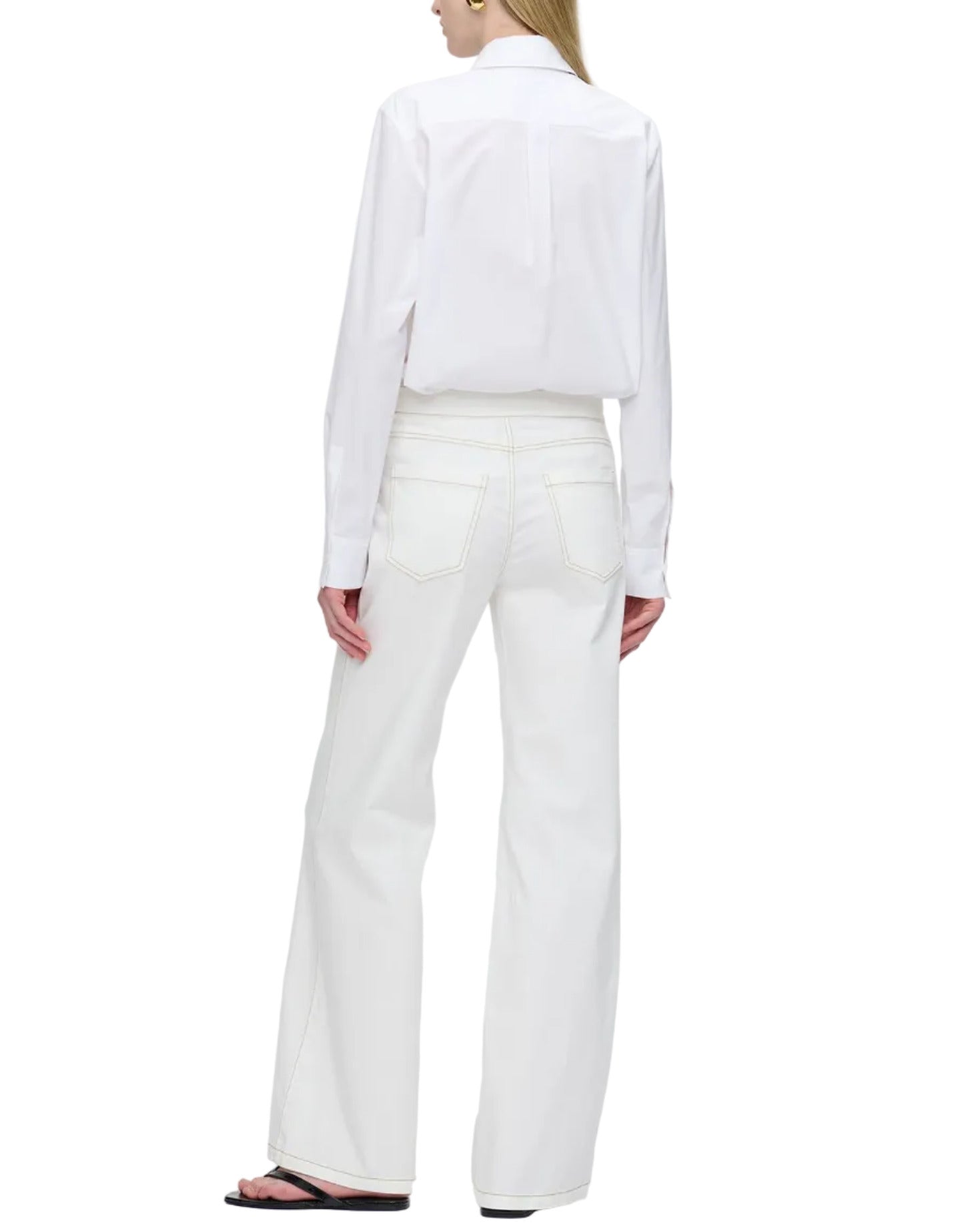 A person stands facing away, wearing a white long-sleeve shirt and Callas Milano's Jane Pant—high rise, wide-leg organic cotton jeans with back pockets—paired with black sandals.