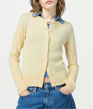 A person wears the pale yellow Jane Cardigan by Guest in Residence over a blue collared shirt with blue jeans, standing hands-in-pockets against a plain background.