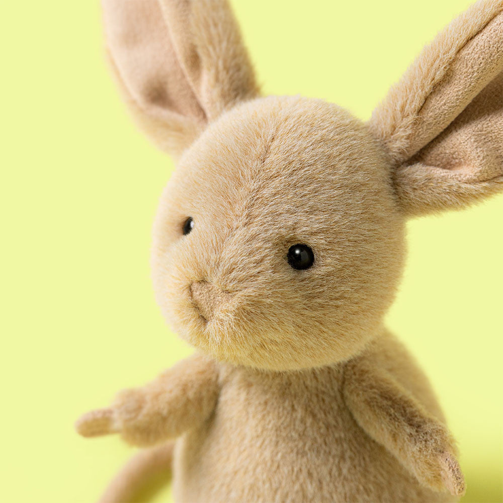 A close-up of Jellycat’s Jerboa plush—a light brown rabbit with large ears and black button eyes—set against a pale yellow background.