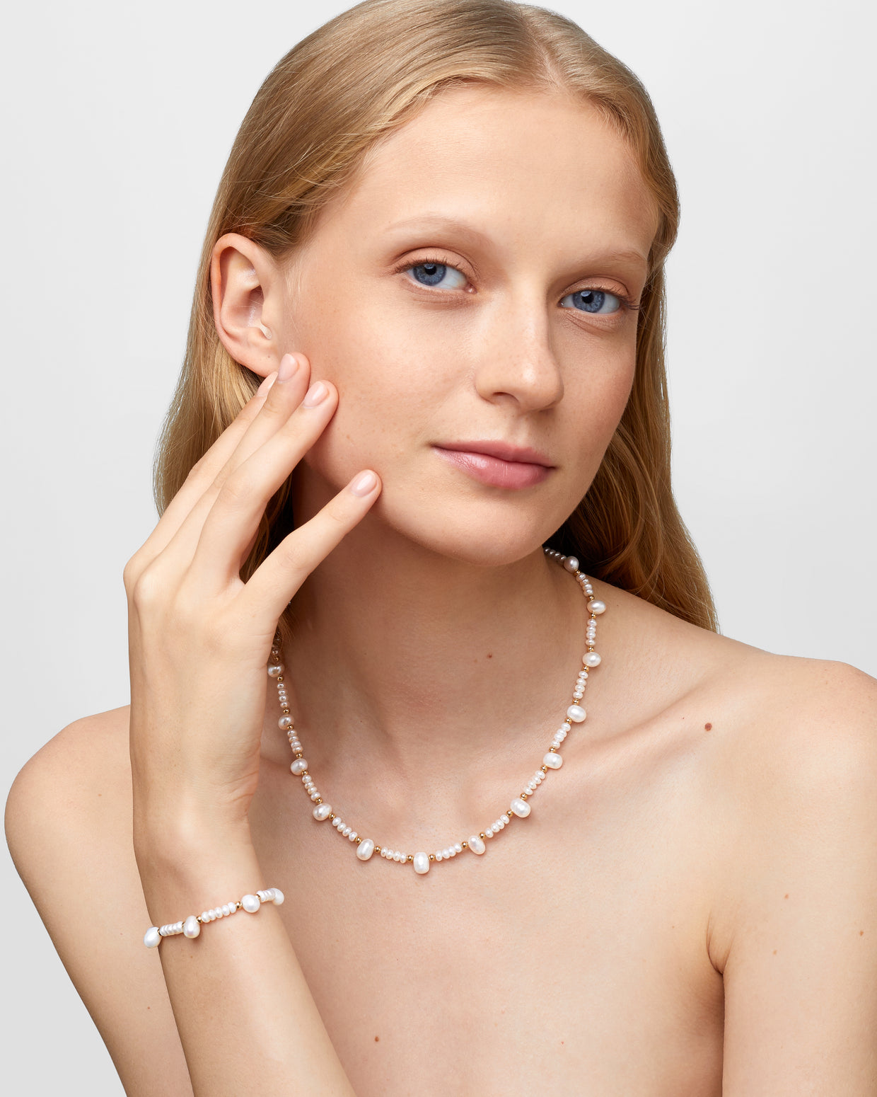 A person with long hair wears a Jia Jia Ocean Pearl Gold Bead Bracelet and necklace, both elegantly crafted from freshwater pearls and 14-karat gold, touching their face gently with one hand.