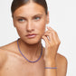 A woman with light skin and light brown hair wears a purple beaded Jia Jia Birthstone Bracelet and matching necklace, lightly touching her face with her right hand against a plain white background.