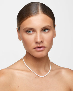 A person with a light skin tone and slicked-back hair wears the Jia Jia Mini Pearl Necklace by Jia Jia, gazing slightly to the side against a plain background.