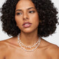 A woman with curly hair is wearing the Jia Jia Ocean Pearl Gold Bead Double Long Necklace, crafted from double-stranded freshwater pearls, as she looks to the side against a plain background.