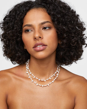 A woman with curly hair is wearing the Jia Jia Ocean Pearl Gold Bead Double Long Necklace, crafted from double-stranded freshwater pearls, as she looks to the side against a plain background.