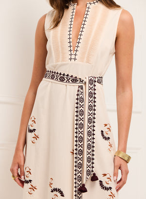 A woman wears the Figue Joan Dress—a sleeveless cream dress with embroidered patterns, deep V-neck, matching embroidered belt—and a gold cuff bracelet.