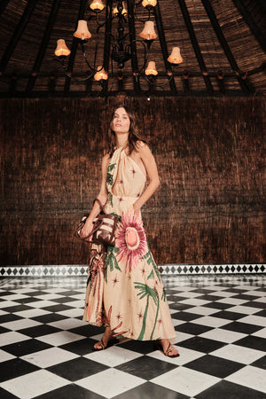 A woman in the Johanna Ortiz Western Elements Ankle Dress holds a brown handbag while standing on a black-and-white checkered floor beneath a chandelier in a thatched-roof room.