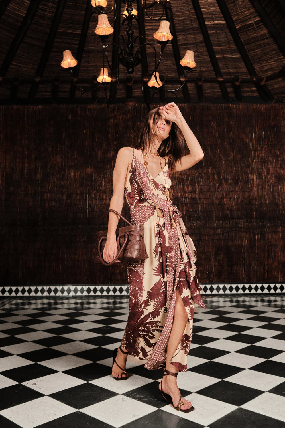 A woman in the Johanna Ortiz Nouvel Arome Maxi Dress and sandals stands on a black-and-white checkered floor under a chandelier, holding a brown bag and touching her head.