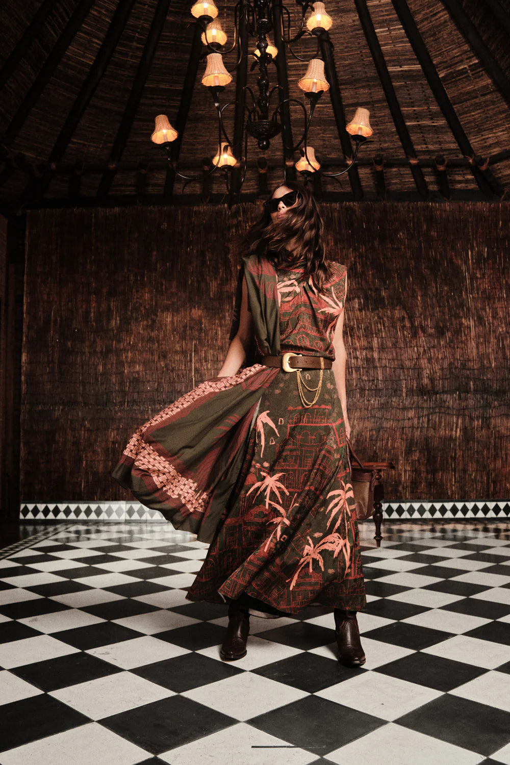 A woman in the Johanna Ortiz Mystic Shore Top and boots stands on a black-and-white checkered floor beneath a chandelier, embodying the brand's signature style in a dim, woven-walled room.