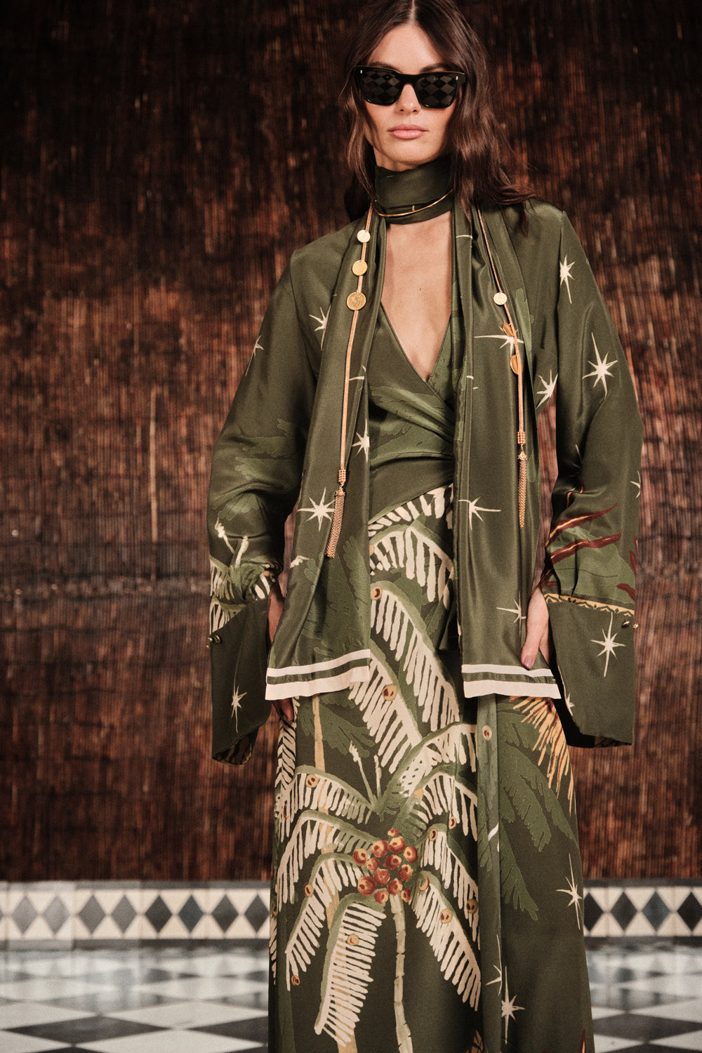 A woman wearing sunglasses and the Johanna Ortiz Eternity Begins Wrap Dress, an olive green silk crêpe de chine ankle-length dress with palm tree and star prints, stands before a brown textured wall and tiled floor.