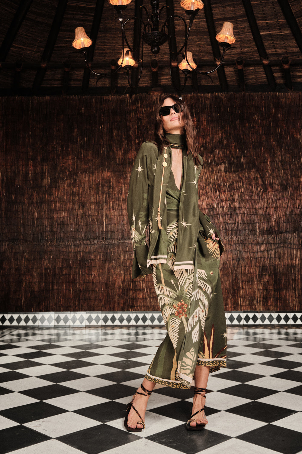 A woman in the Johanna Ortiz Eternity Begins Wrap Dress—an olive green, tropical JO print, silk crêpe de chine, ankle-length gown—wears sunglasses and sandals as she stands on a black-and-white checkered floor beneath a chandelier.