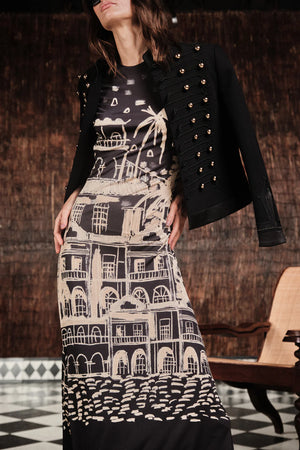 A woman stands indoors near a wooden chair, wearing the Johanna Ortiz Cardinal Direction Ankle Dress—a beige and black architectural print dress—paired with a black jacket featuring decorative buttons.