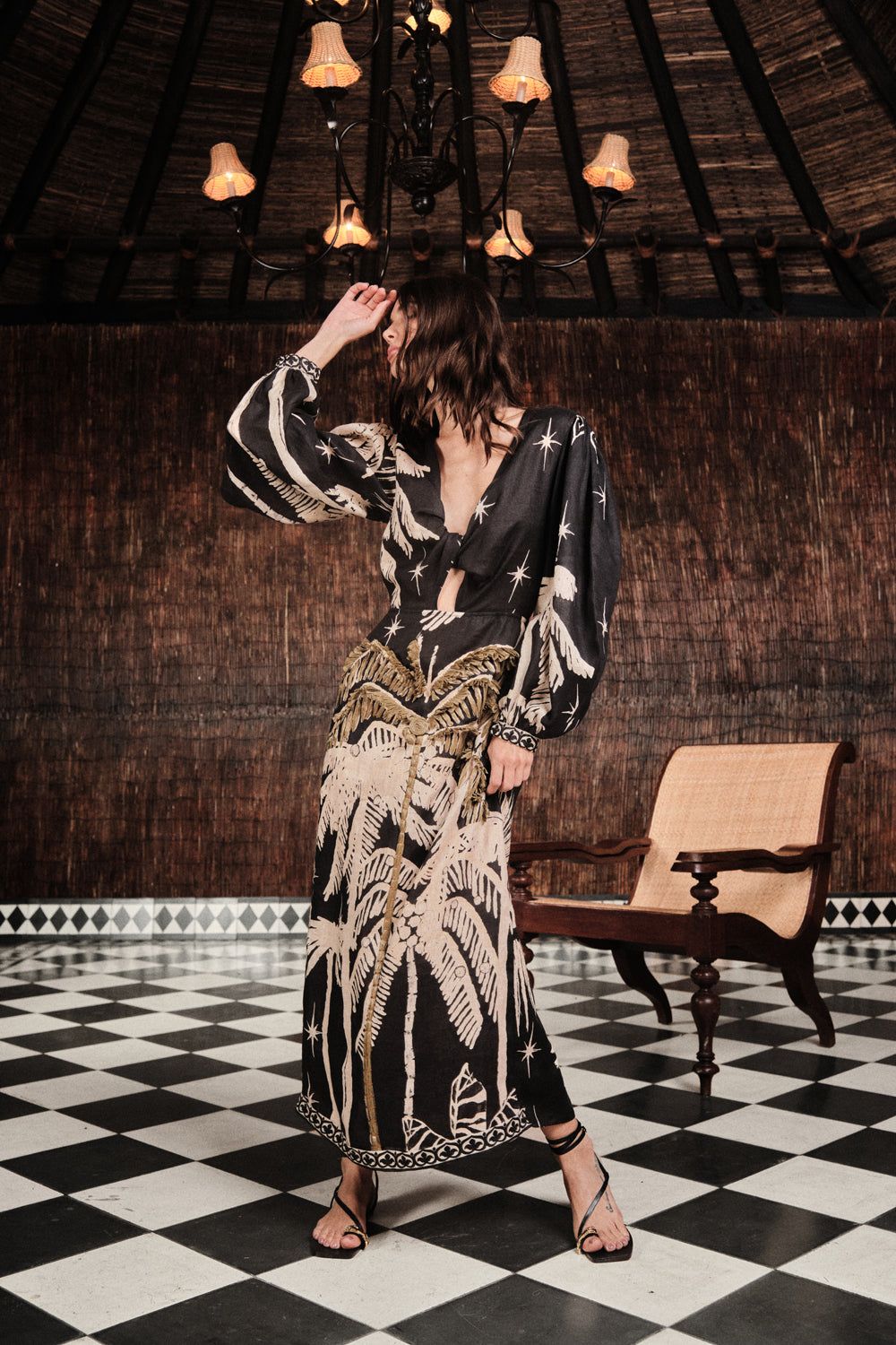 A woman in the Pradera Geometrica Ankle Dress by Johanna Ortiz, featuring beige palm motifs, poses gracefully on a checkered floor, evoking effortless resort elegance.