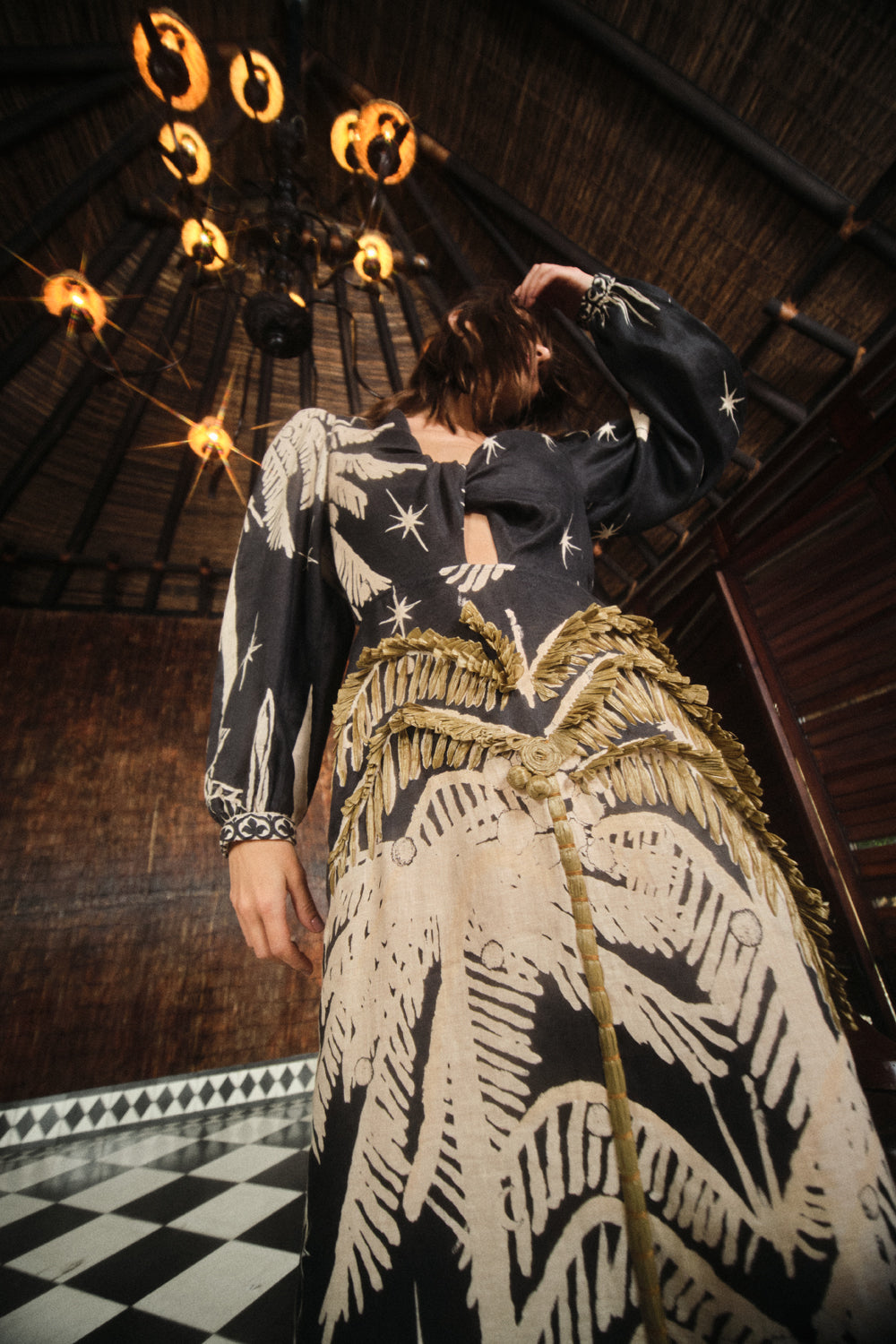A person in the Pradera Geometrica Ankle Dress by Johanna Ortiz stands indoors beneath a circular light fixture, with one arm raised and their face partially obscured.