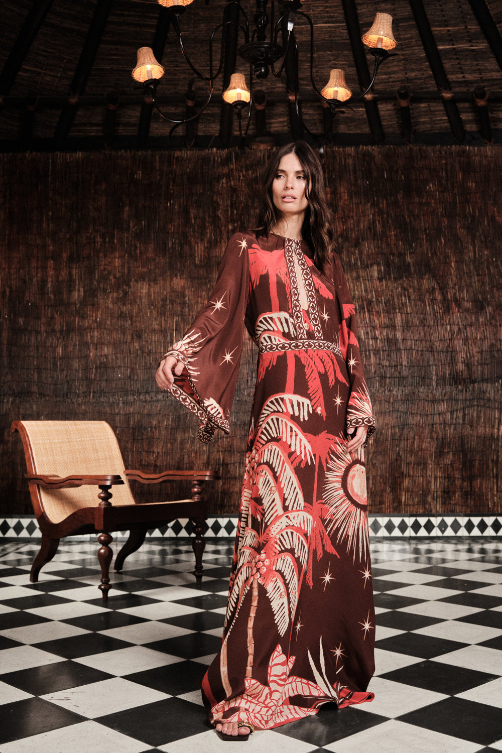 A woman stands on a black-and-white checkered floor wearing the Johanna Ortiz Leyenda Del Tiempo Maxi Dress, featuring palm and star motifs, with a wicker chair and wooden wall in the background.
