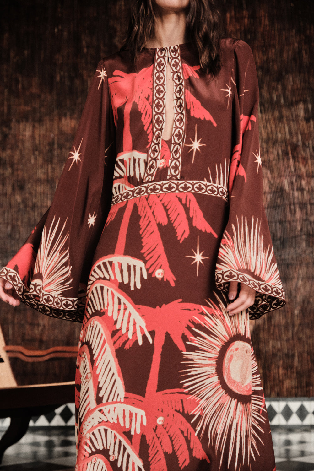 A woman wears the Johanna Ortiz Leyenda Del Tiempo Maxi Dress, featuring long sleeves and a red and beige tropical palm tree and sun pattern on a dark background.