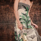 A woman wears the Johanna Ortiz Simbologia Floral Maxi Dress, cream-colored with green and black tropical motifs, holding a small gold purse on a black-and-white tiled floor against a brown textured background.