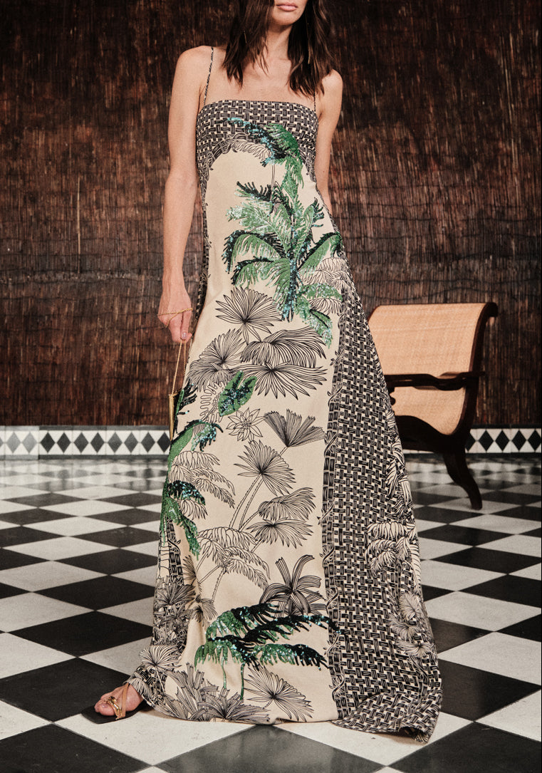 A woman wears the Johanna Ortiz Simbologia Floral Maxi Dress, featuring strapless styling with botanical embroidery and geometric patterns, as she stands on a black-and-white checkered floor.