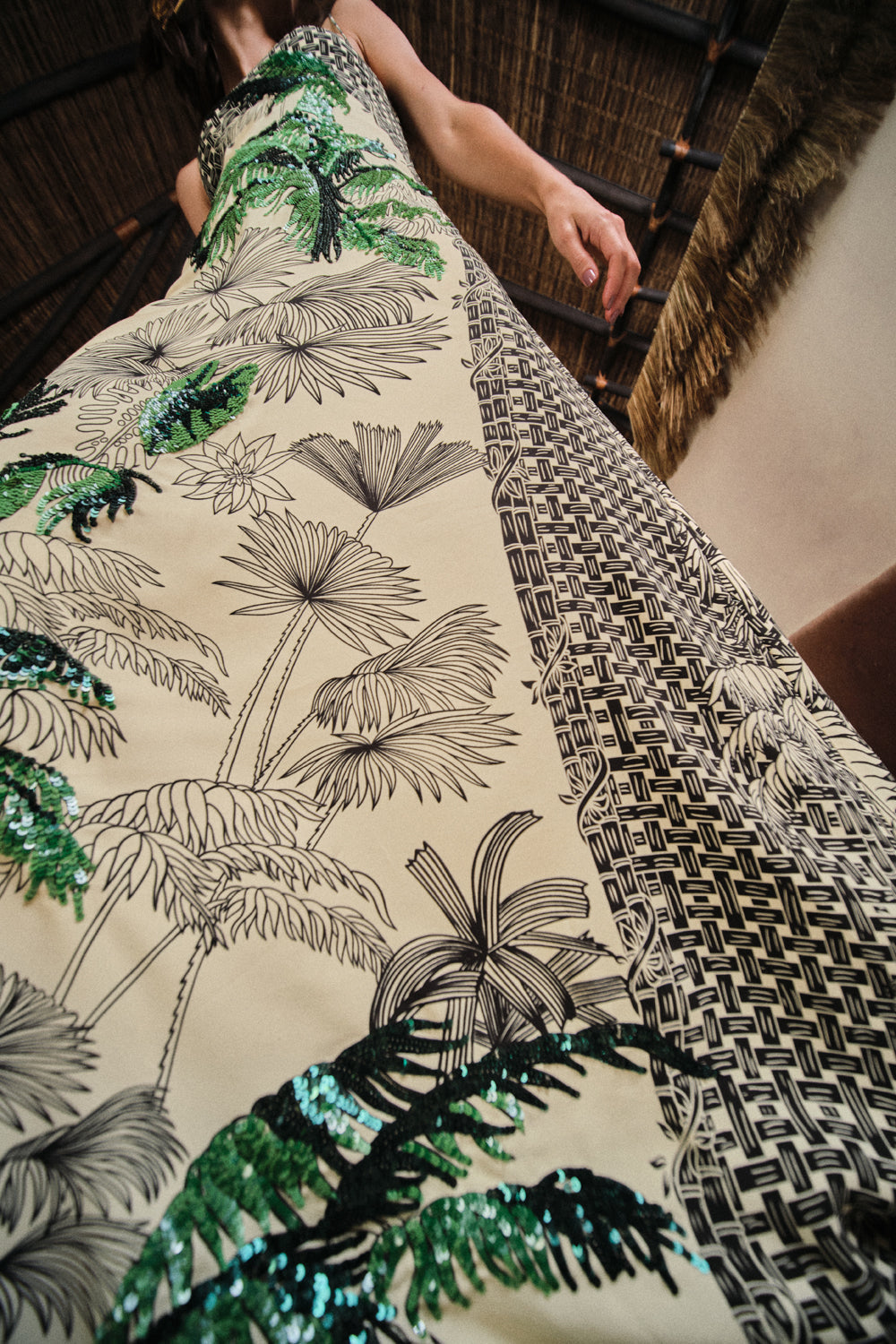 A person wearing the Johanna Ortiz Simbologia Floral Maxi Dress, featuring green sequined leaves, black line palm motifs, and geometric shapes, photographed from a low angle indoors.