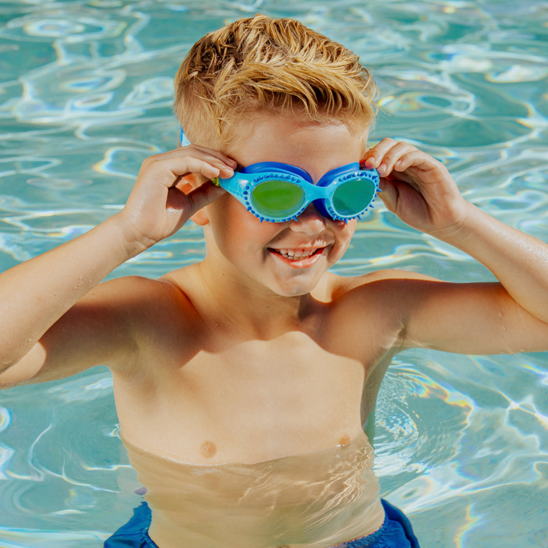 A young boy in a pool smiles as he adjusts his blue Bling2O Kids' Jurassic Dive Goggles.