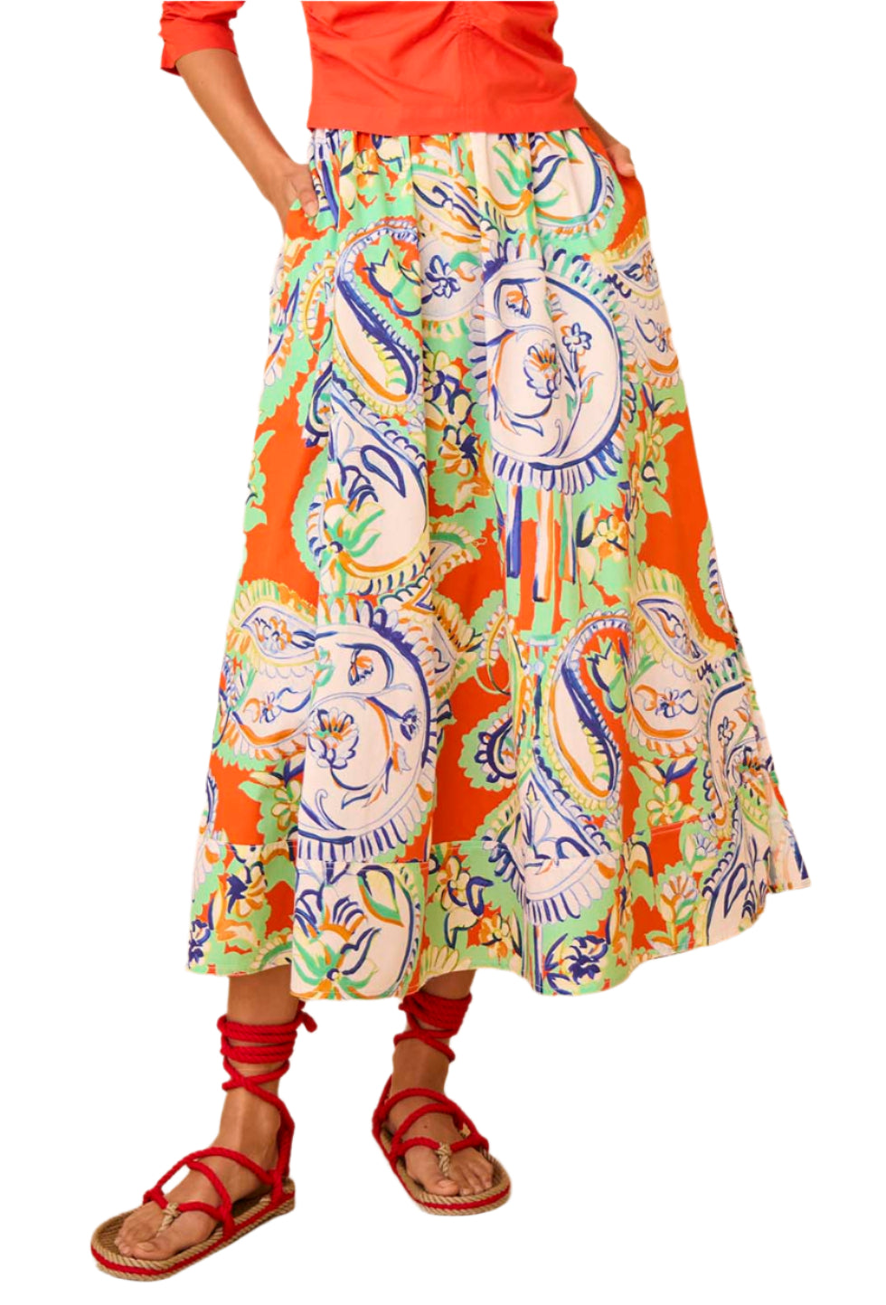 A person is wearing the Hunter Bell Tatum Skirt from Hunter Bell NYC, featuring a vibrant floral pattern on an orange background and an elastic waistband, paired with a red top and matching red sandals.