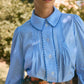 A person stands outdoors wearing the Hunter Bell NYC James Shirt, a light blue Saltwater Check button-down blouse with scalloped collar and buttons, paired with a floral skirt.
