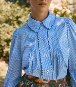 A person stands outdoors wearing the Hunter Bell NYC James Shirt, a light blue Saltwater Check button-down blouse with scalloped collar and buttons, paired with a floral skirt.