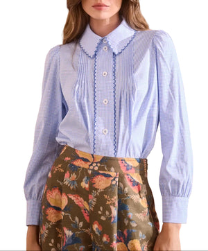 A woman wearing the James Shirt by Hunter Bell NYC—a light blue gingham blouse with scalloped trim and white buttons—paired with high-waisted, floral-patterned pants.